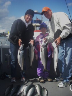 Two adults and a child on a boat proudly display freshly caught fish, holding them up for the camera. Additional fish are arranged on the boat's floor. Bright, cloudy sky in the background.
