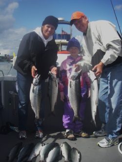 Two adults and a child on a boat proudly display freshly caught fish, holding them up for the camera. Additional fish are arranged on the boat's floor. Bright, cloudy sky in the background.