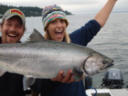 Two people on a boat happily holding a large fish they caught; the water and shoreline are visible in the background.