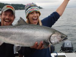 Two people on a boat happily holding a large fish they caught; the water and shoreline are visible in the background.