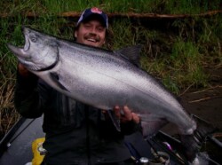 A person in outdoor clothing holds a large fish with a smile against a backdrop of grass and a log.