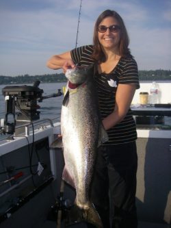A woman in sunglasses and a striped shirt stands on a boat holding a large fish. The background shows a body of water and trees on a distant shore.